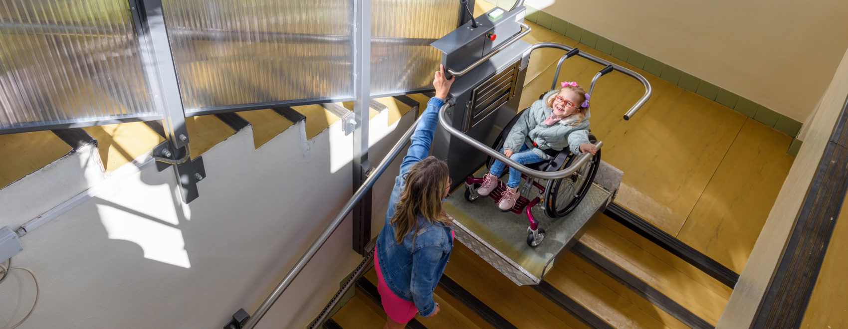 Mother with a young child living with cerebral palsy using electric wheelchair lift to access public building. Special lifting platform for wheelchair users. Disability stairs lift facility.